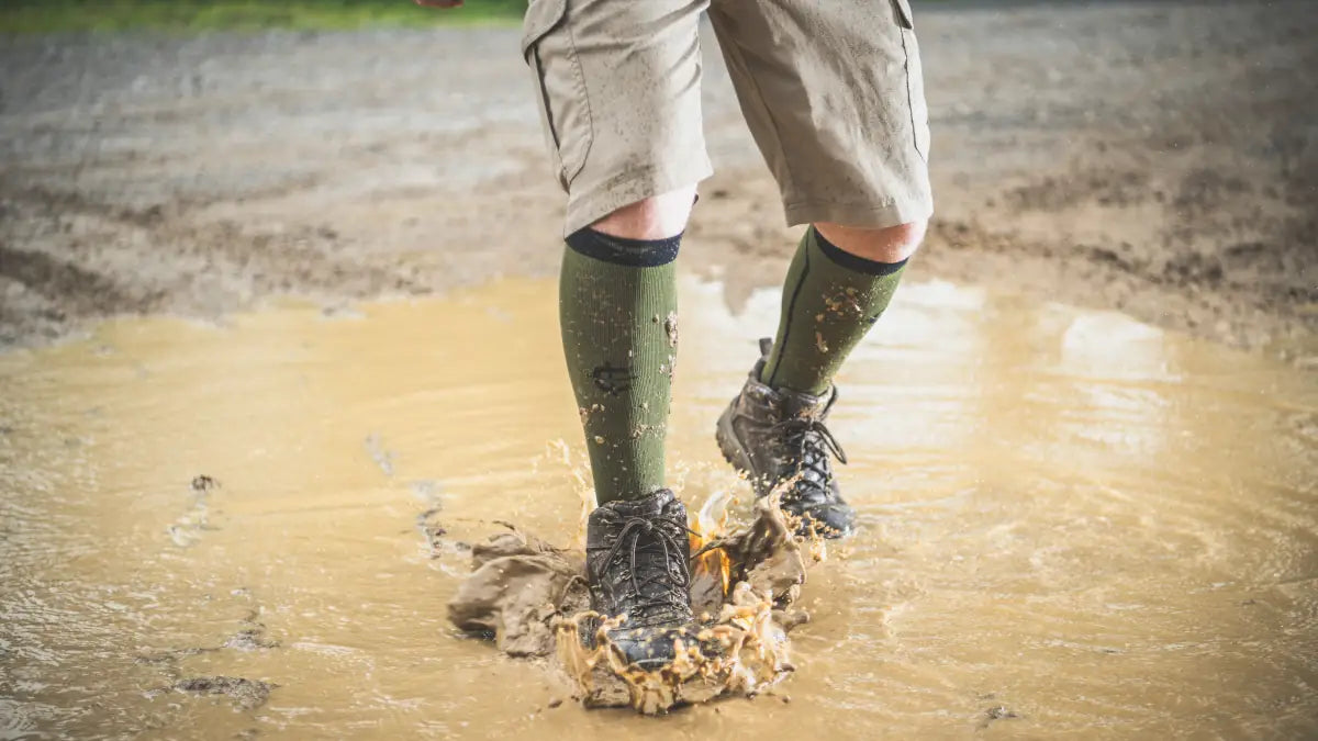 Over-the-calf outdoor socks and laced boots splashing through mud, one of the many kinds of terrain Covert Threads outdoor socks are made for.