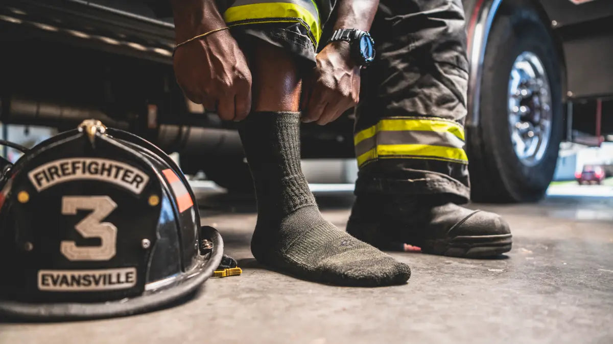 A firefighter putting on Covert Threads work socks by a fire helmet and boots, one of many work sock styles for first responders and the trades